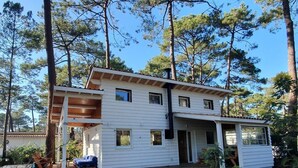 A two-story white house with a wooden deck and a covered porch, surrounded by tall pine trees.