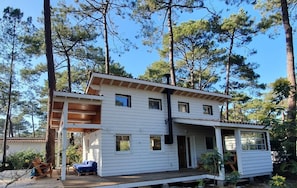 Uma casa branca de dois andares com varanda de madeira e alpendre coberto, cercada por altas árvores de pinheiro.