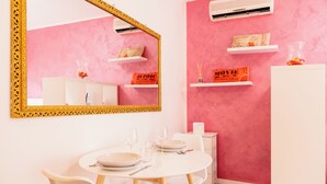 Un coin repas avec une table ronde, des chaises blanches et un miroir encadré de doré. On y trouve des étagères blanches ornées d’objets décoratifs et un meuble blanc.