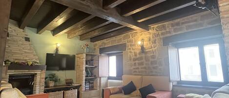 A cozy living room with a fireplace, stone walls, and wooden beams. There are sofas and a television mounted on the wall.