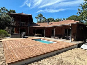 A wooden deck with a pool, a covered patio area, and a two-story building with a balcony. - Wooden villa surrounded by nature + heated pool + spa + ocean pools in Soulac (Le Verdon-sur-Mer)