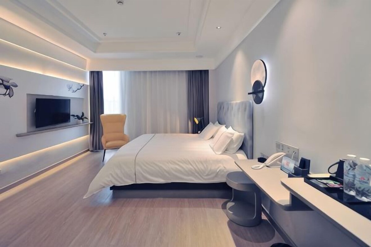 A modern hotel room with a large bed, a flat-screen TV mounted on the wall, a desk with a phone, and a chair.