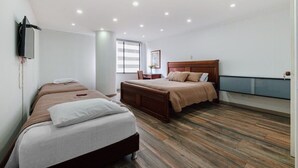 Ein Hotelzimmer mit zwei Betten, einem Holzkopfende, einem Nachttisch, einem an der Wand montierten Fernseher und einem Fenster mit Jalousien.
