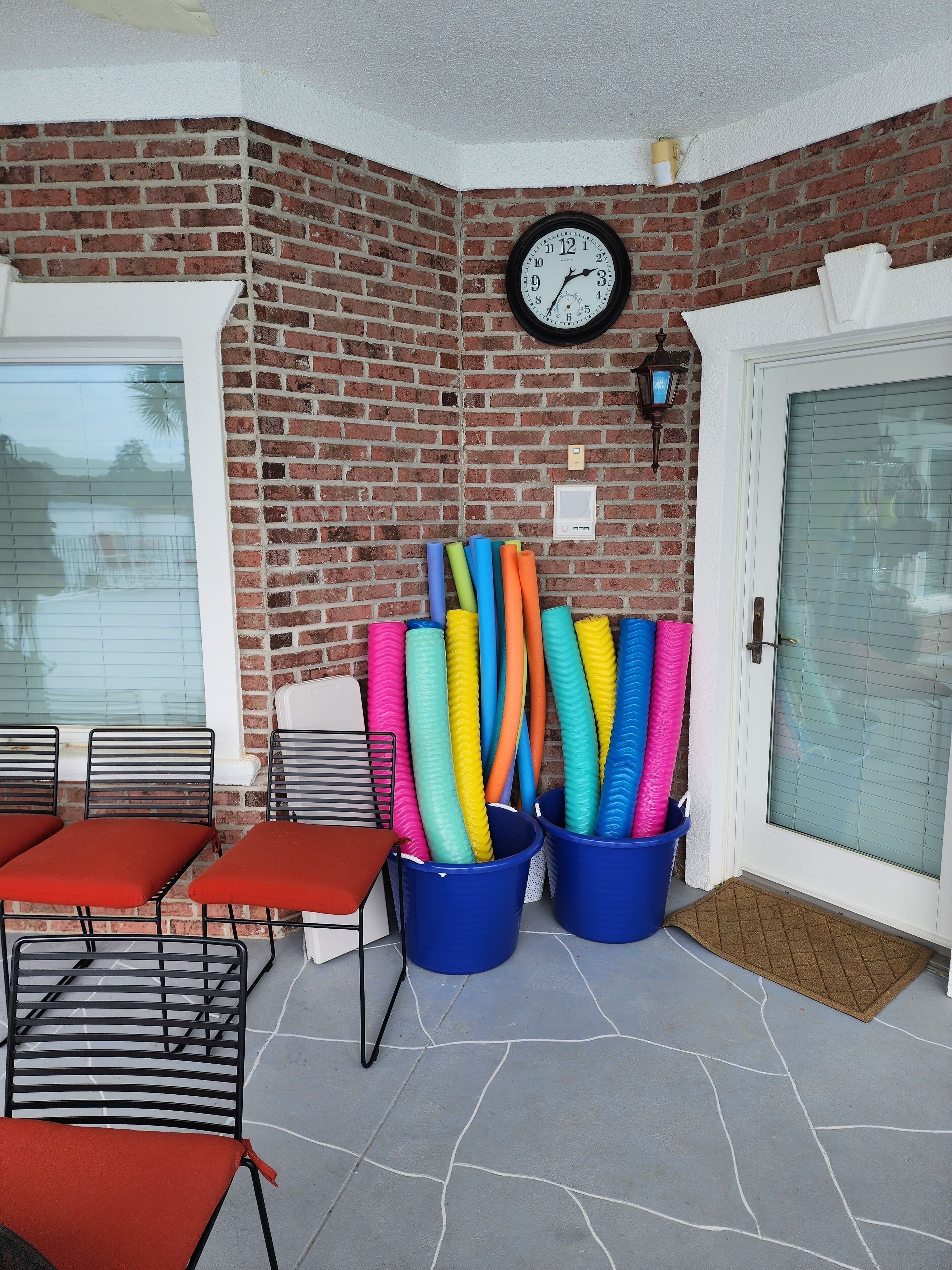 Ein Patio mit einer Ziegelmauer, einer Uhr und bunten Matten in blauen Eimern.