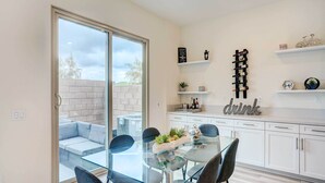 Une cuisine moderne avec une table à manger en verre, des meubles blancs et une porte-fenêtre coulissante donnant sur un espace de détente extérieur.