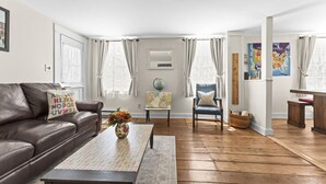 A living room with a brown leather sofa, a wooden coffee table with a floral vase, a blue armchair, and a wooden side table. - Historic Waterfront School House (Groton)