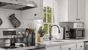 A modern kitchen with stainless steel appliances, white cabinetry, and a window with a view of greenery. - Historic Waterfront School House (Groton)