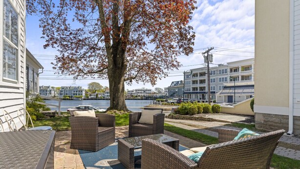 A patio area with wicker furniture, a large tree, and a view of a waterfront and buildings in the background. - Historic Waterfront School House (Groton)