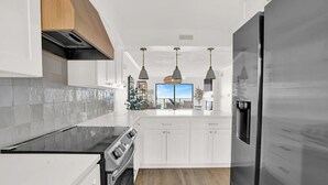 Una cocina moderna con armarios blancos, encimera negra y electrodomésticos de acero inoxidable. Hay luces colgantes sobre la isla y vistas al exterior a través de un ventanal grande.