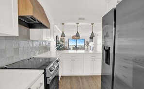 A modern kitchen with white cabinetry, a black countertop, and stainless steel appliances. There are pendant lights above the island and a view of the outdoors through a large window.