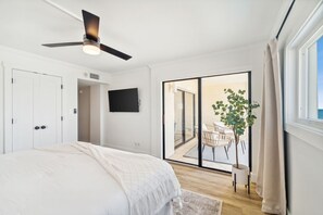 Un dormitorio con una cama, ventilador de techo, televisor, puerta corrediza de vidrio a un balcón y una planta en maceta.