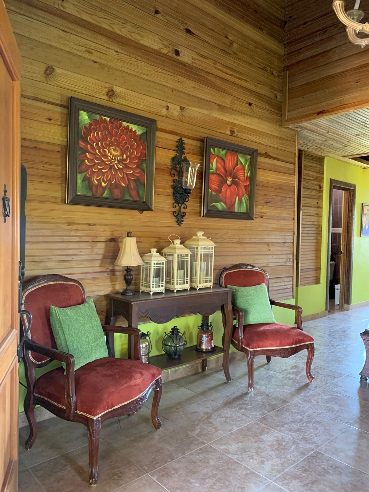 A wooden-paneled room with two red upholstered chairs, a small table with a lamp, lanterns, and wall-mounted flower paintings.