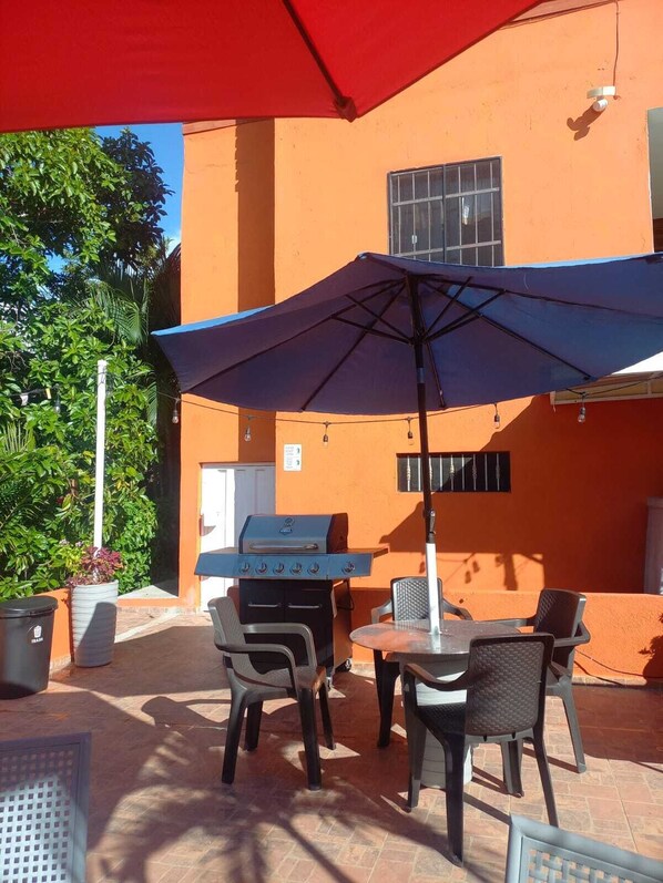 Eine Terrasse im Freien mit einem Grill, zwei Stühlen und einem Tisch unter einem roten Sonnenschirm.