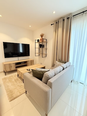 A modern living room with a flat-screen TV, a wooden coffee table, a grey sofa, and a beige rug. - Comfortable one bedroom Studio Apartment @ The Lennox Airport Residential Accra (Accra)