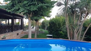 Una piscina con muelle de madera y sillones de descanso, rodeada de árboles y un área de patio techada.
