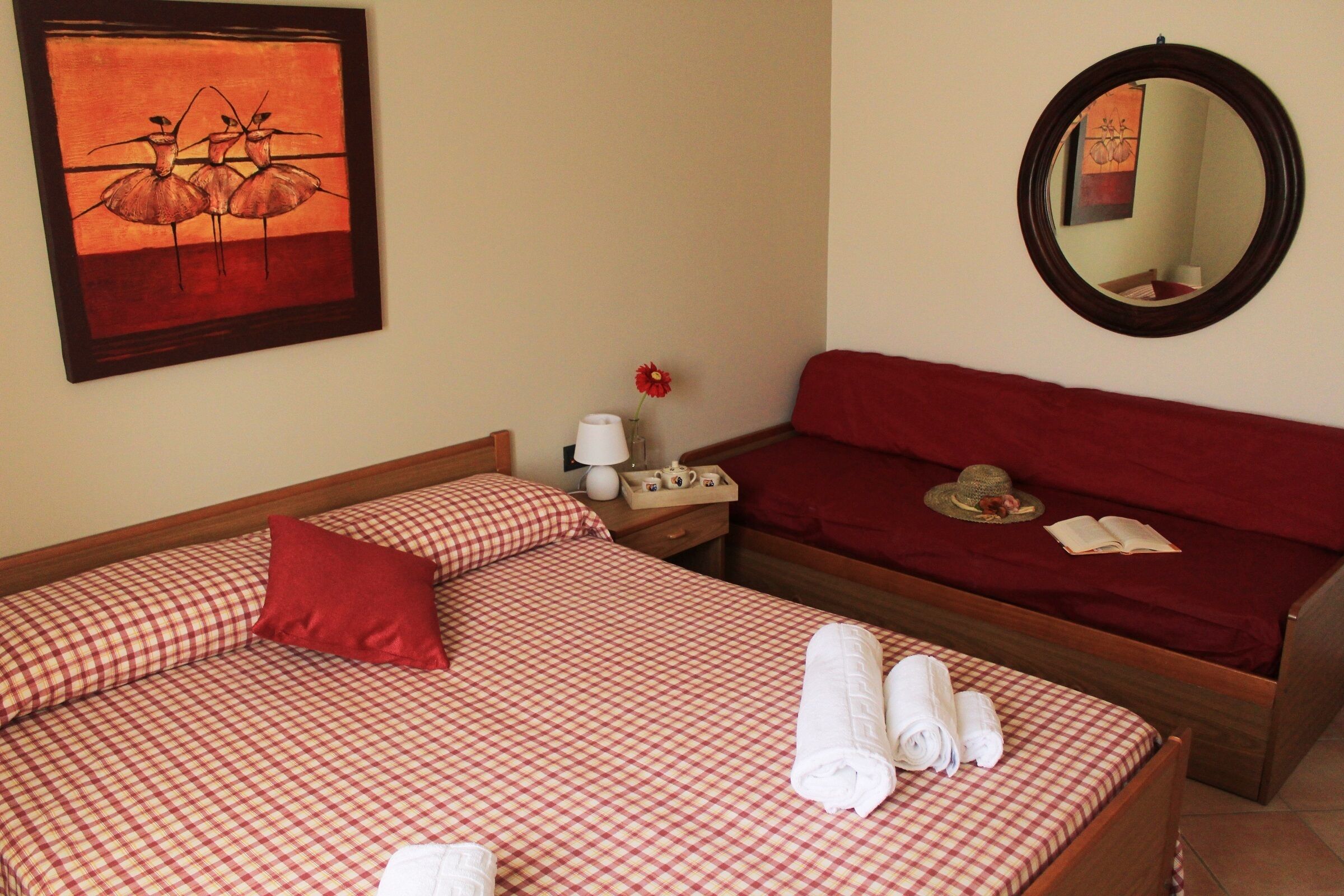 A hotel room with two beds, checkered bedding, a red headboard, a mirror, a painting, a vase with a flower, and a nightstand with a lamp and a cup.