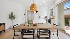 A modern kitchen with white cabinets, a central island, and a dining area with a wooden table and chairs. - Desert mountain views w/ pool, fire pit & hot tub (La Quinta)