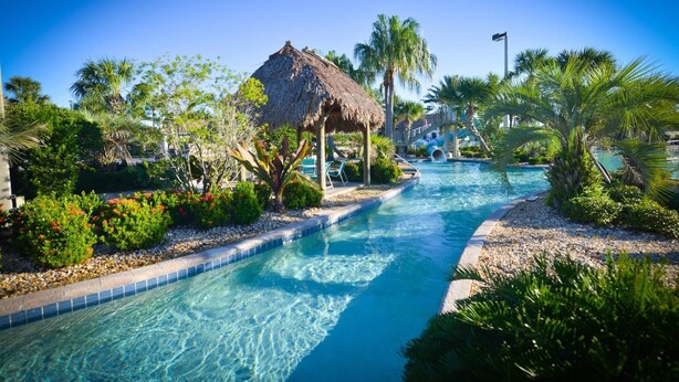 A clear pool with a thatched-roof structure, surrounded by tropical plants and palm trees. - Mickey's Mansion Luxury Themed Pool Home, Arcade, Theater (Four Corners)