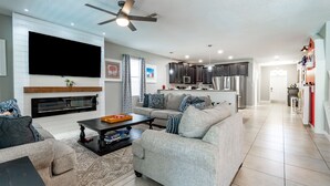 A modern living room with a sofa, coffee table, and TV. There is a fireplace, kitchen area with cabinets and appliances, and a ceiling fan. - Mickey's Mansion Luxury Themed Pool Home, Arcade, Theater (Four Corners)