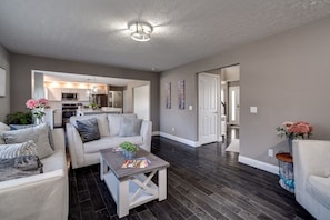 A spacious living room with a dark wood floor, white sofas, and a coffee table. A kitchen is visible in the background with modern appliances and pendant lighting. - 6BR Home w/ Pool - Spooky Nook/MiamiU/KingsIsland (Hamilton)