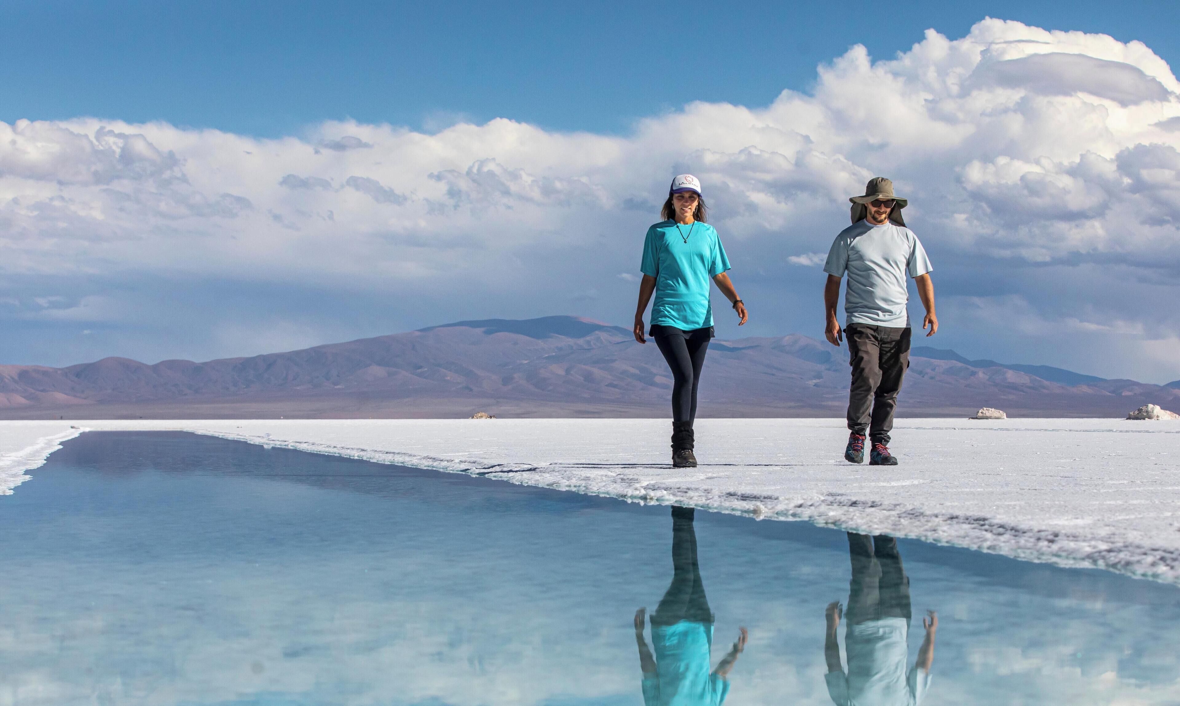 Two people walking on a reflective surface with mountains in the background.