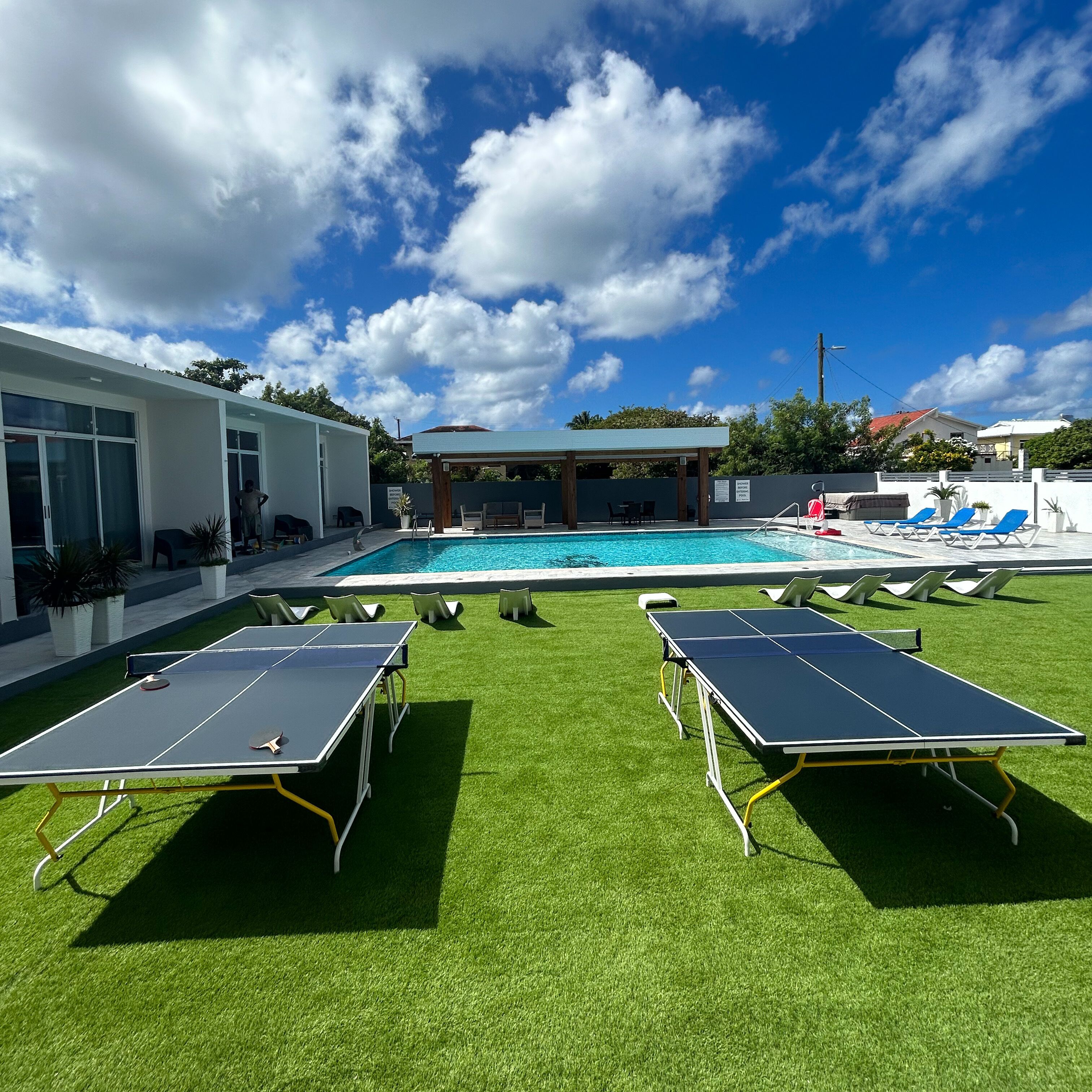 Zona de piscina con dos mesas de ping pong, un área de estar cubierta y una piscina.