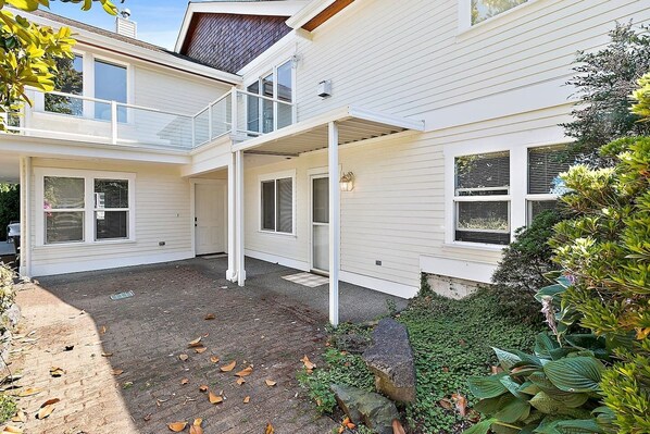 A two-story house with a covered patio, a front door, and a small garden area with plants and a stone bench. - Cozy Old Town condo + Garage + Patio Steps to Ruston Way! (Tacoma)
