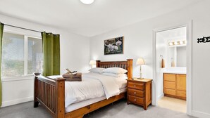 A bedroom with a wooden bed, a nightstand, a bedside table, a window with blinds, a green curtain, a painting on the wall, and a bathroom visible through an open door. - Cozy Old Town condo + Garage + Patio Steps to Ruston Way! (Tacoma)