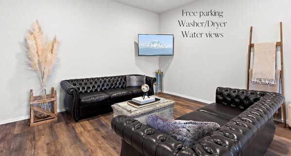 A modern living room with a black leather sofa, a wooden coffee table, and a mounted TV. Amenities include free parking, a washer/dryer, and water views.