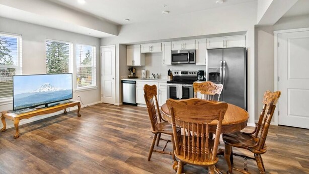 A modern kitchen with white cabinets, stainless steel appliances, and a dining area with wooden chairs and a television displaying a mountain view. - Quiet condo Steps to Tacoma Dome~Bay & City VIEWS! (Tacoma)