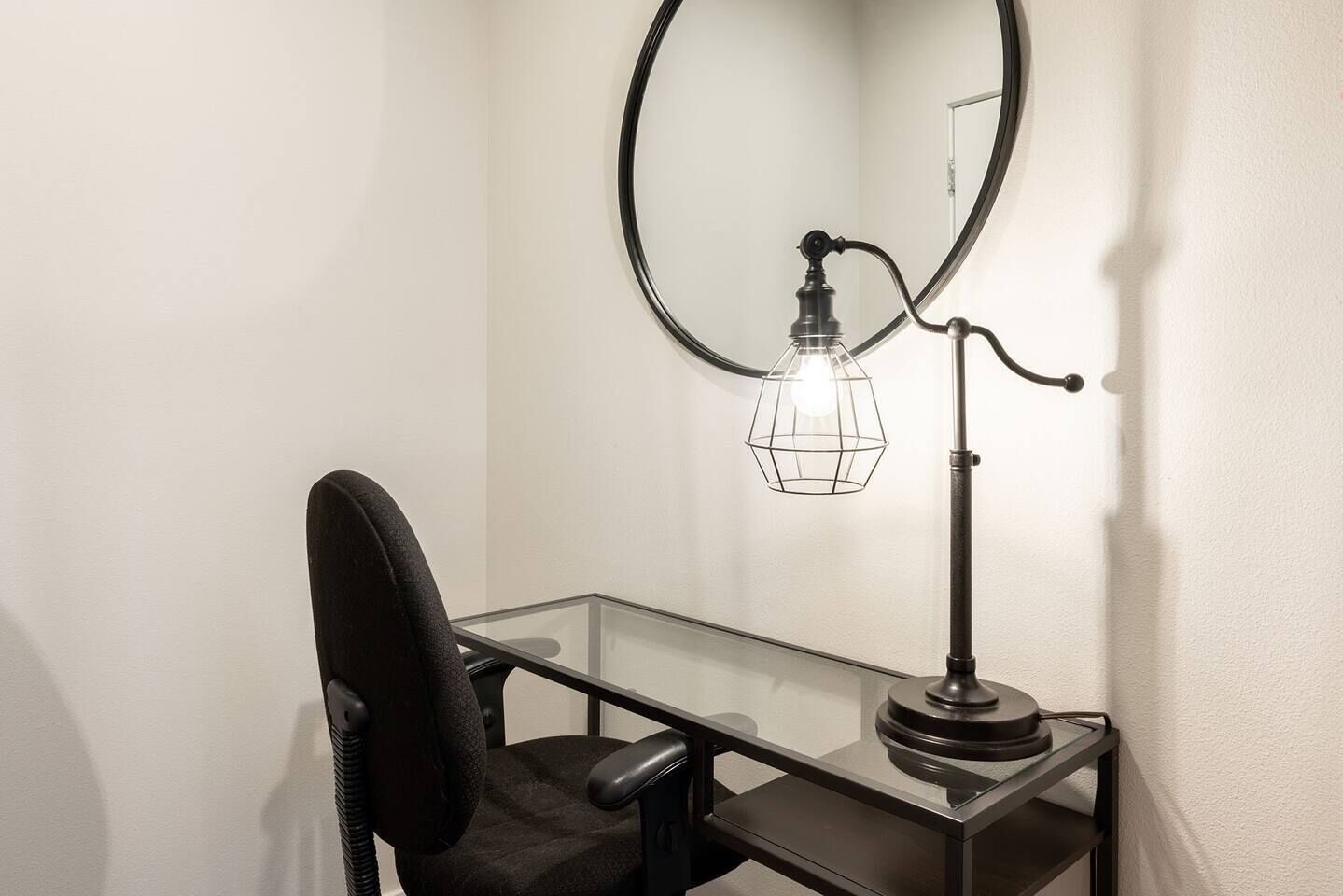 Un espacio de oficina moderno con un escritorio de cristal, una silla de oficina negra y una lámpara de escritorio única con base circular y pantalla de entramado de alambre.