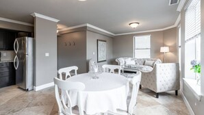 A modern kitchen with a stainless steel refrigerator, a dining area with a round table and white chairs, and a living room with a sofa and a lamp.