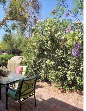 Relax in a serene garden setting with a patio featuring a table and chairs surrounded by lush greenery and flowering plants.