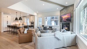 Amplia sala de estar con sofá, una mesa de centro y chimenea. Se observa una cocina con electrodomésticos modernos y un conjunto de comedor. La habitación cuenta con grandes ventanales que ofrecen luz natural y vistas al exterior.