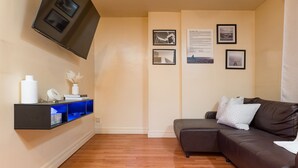 Un salón con un sofá de cuero marrón, un televisor de pantalla plana montado en la pared, una estantería con objetos decorativos e imágenes enmarcadas.