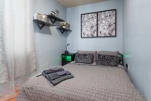 Ein Schlafzimmer mit einem Bett, Kissen, einem Nachttisch, einer an der Wand befestigten Ablage mit Dekoration und einem gerahmten floralen Kunstwerk.