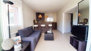 A modern living room with a grey sofa, a coffee table, a TV, and a kitchen area with a counter and pendant lights.