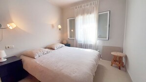 A bedroom with a large bed, two bedside lamps, a small stool, and a window with sheer curtains.