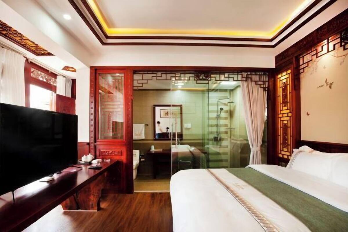 A hotel room with a bed, a desk, a television, and a bathroom visible through a glass door.