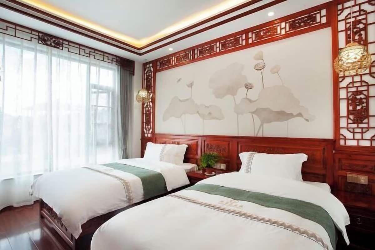 A hotel room with two beds, a large window with curtains, a decorative wall with a floral painting, and a traditional Chinese-style wall panel.