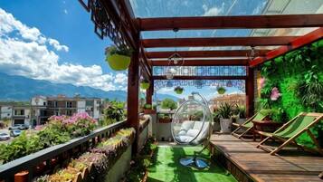 Un balcón con suelo de madera, plantas en macetas, una silla colgante y vistas a edificios y montañas.