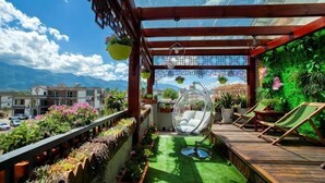A balcony with a wooden floor, hanging plants, a hanging chair, and a view of buildings and mountains. - Yamingyuan Inn (Dali City)