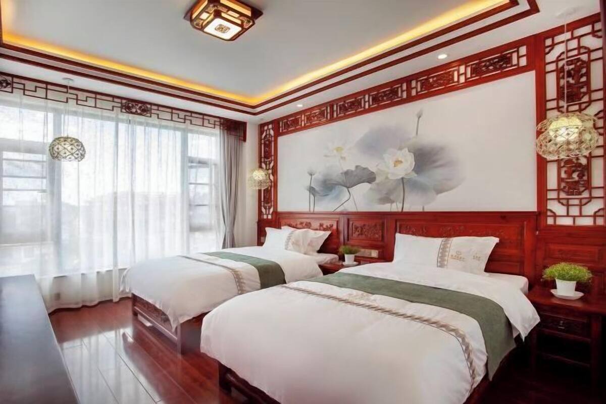 A hotel room with two beds, a large window with sheer curtains, a decorative wall with traditional patterns, and a ceiling light fixture.
