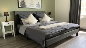 Un dormitorio ordenado con una cama, dos mesitas de noche, una obra de arte enmarcada y una ventana con cortinas.