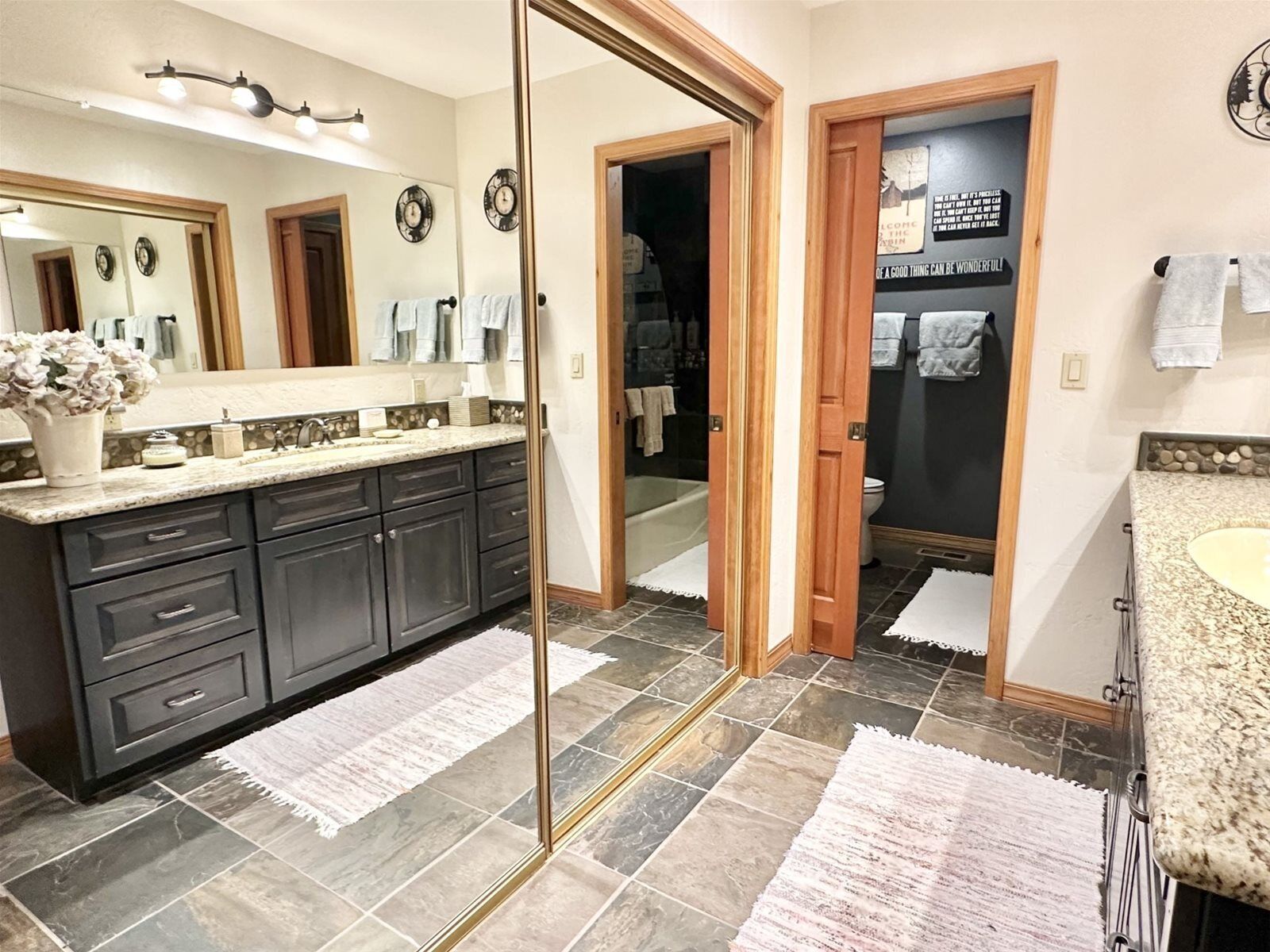 A modern bathroom with dark gray cabinets, a large mirror, and a vanity with a granite countertop. There are two sinks, a shower, and a toilet visible.