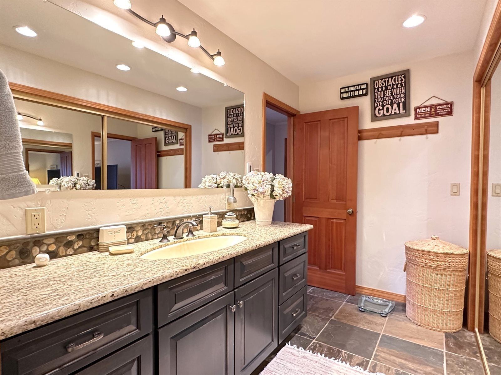 A bathroom with a granite countertop, dark cabinetry, a large mirror, and motivational wall signs.