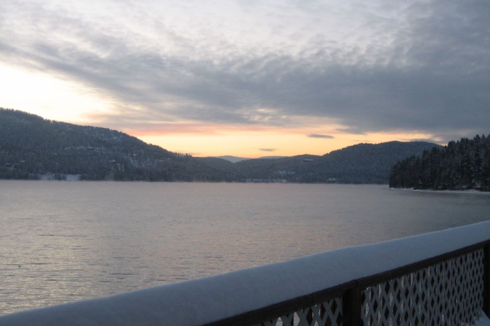 A lake with snow-covered hills and a wooden railing in the foreground.
