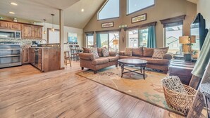 A spacious living area with a kitchen, wooden flooring, and large windows.
