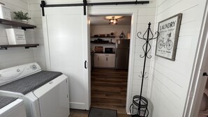 A laundry room with a washing machine, dryer, and a rack with folded clothes. There is a door leading to a kitchen area with a refrigerator, microwave, and a hanging light fixture. - 7 mi to Grand Park, 1 mi to Mustard Seed, 5 min to downtown Noblesville! (Noblesville)