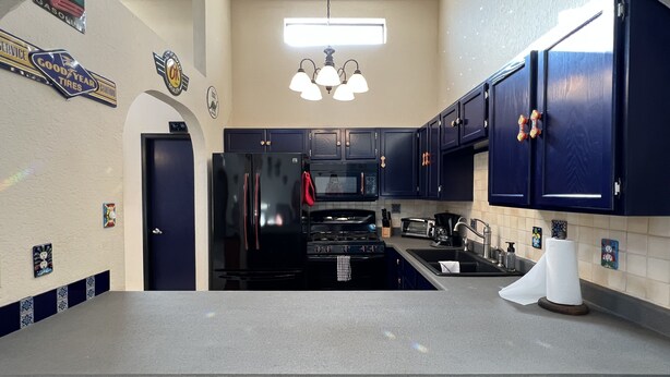 Una cocina con armarios azul oscuro, un refrigerador negro, una estufa y un microondas. Hay letreros decorativos en la pared y una ventana sobre el fregadero.